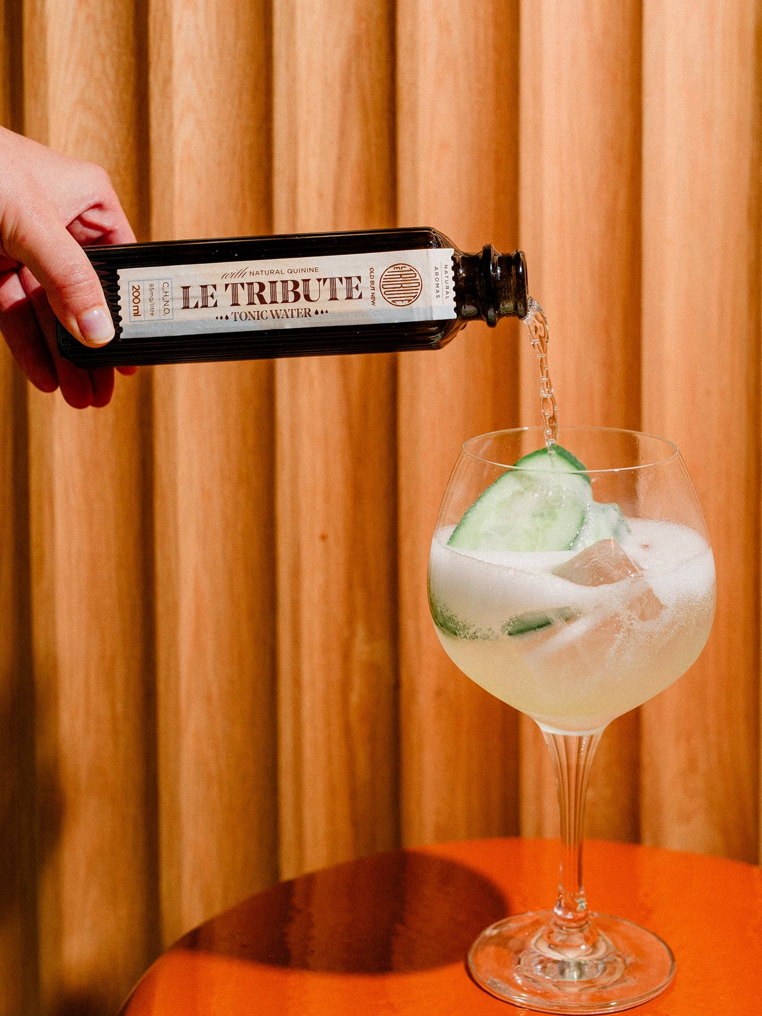 Hand pouring LE Tribute Tonic Water into a glass with ice and cucumber for a refreshing cocktail
