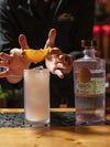 Bartender garnishing a cocktail with orange slice, featuring LE Tribute spirit bottle on the bar