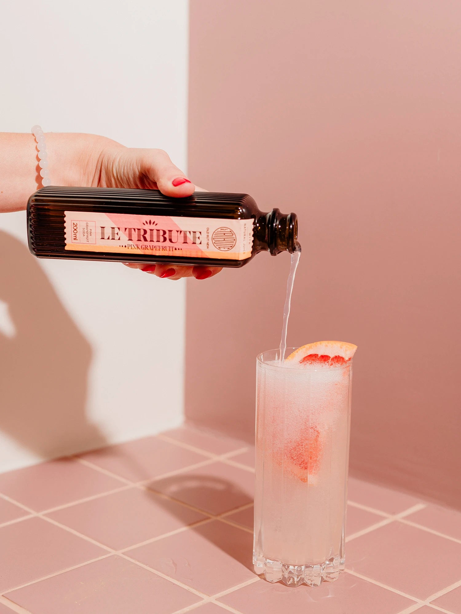 Hand pouring LE Tribute Pink Grapefruit Soda into a glass with ice and grapefruit for a refreshing cocktail